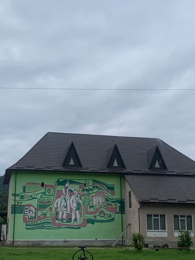 The image displays a two-story building located in Tatariv, Ukraine, under an overcast sky. The building features a dark brown, multi-pitched roof with three triangular dormer windows. The left and central portion of the building's facade is painted lime green and features a large, folk art-style mural. The mural, rendered in green, red, and white hues, depicts a central group of three figures in traditional attire, one playing a wind instrument and another a drum, surrounded by abstracted representations of houses, landscapes, and mechanical elements. The right portion of the building's facade is light gray stucco with two ground-level sets of rectangular windows. A light-colored wooden ladder is propped against the lime green section of the building's wall. In the immediate foreground, green grass is visible, along with a dark, metallic, curvilinear object partially visible in the lower center. Small green shrubs are present near the base of the building on the right. A single, thin utility wire spans horizontally across the upper third of the frame. Green hills are visible in the background, particularly on the left side.