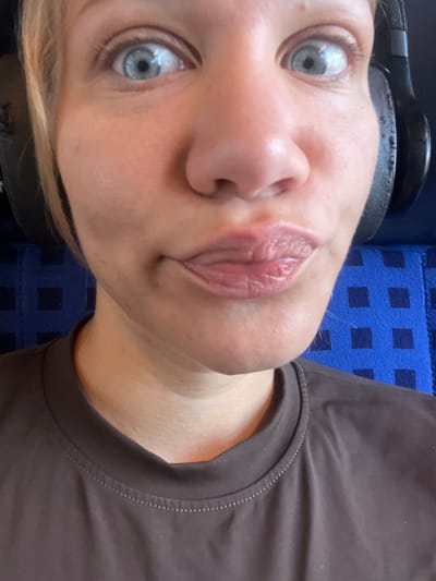 The image is a close-up, selfie-style photograph of a person wearing headphones and a dark brown t-shirt. The person has blue eyes and their lips are pursed. They are sitting in front of a blue, checkered surface, possibly a seat on public transport. The setting appears to be inside a train car in Ampfing, Germany.