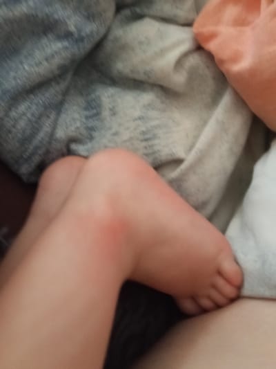 The image presents a close-up view of a person's legs, likely a child's, resting indoors in Teykovo, Russia. The legs are bare, and one knee exhibits a slight reddish discoloration. Small, soft feet are visible at the end of the limbs, suggesting a young individual. The person appears to be in a relaxed, possibly sleeping or resting, position. Surrounding the legs are several fabric items, including a rumpled blue and grey patterned blanket, an off-white blanket, and a peach-colored textile. The background immediately behind the legs is dark. No specific activity, emotions, time of day, or weather conditions are discernible from the photograph. There is no visible text.