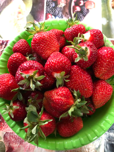 A bright green plastic bowl overflowing with ripe, red strawberries is presented. The strawberries are varied in size and shape, with visible seeds on their surfaces and green stems attached. The bowl sits on a surface that appears to be a tablecloth with a floral or abstract pattern in shades of red and white, and a blurred background shows hints of other images, possibly food-related. The lighting suggests an outdoor or well-lit indoor setting. There are no people visible in the image. The scene does not provide any specific information about the city of Staroe Istomino, Russia.