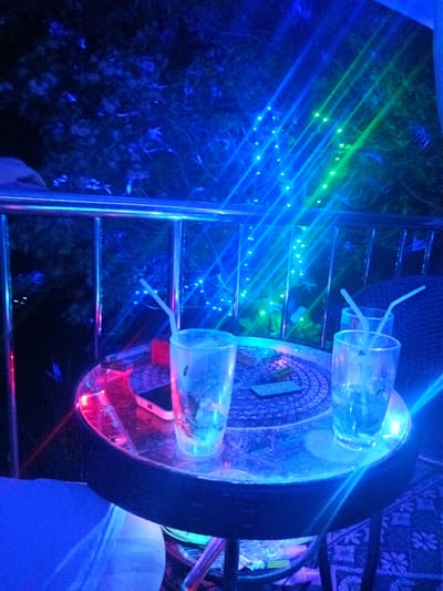 The image depicts a dimly lit outdoor setting, possibly a bar or patio, in Moo 4, Thailand. A small, round table with a dark edge and a mirrored top dominates the foreground. On the table, there are two empty drinking glasses, each containing a white plastic straw. A phone and an unidentifiable object rest on the table as well. The table is placed on a woven, patterned mat. A metal railing runs behind the table, suggesting an elevated location like a balcony. Beyond the railing, trees are visible, illuminated by a combination of blue and green string lights. The entire scene is bathed in blue lighting, creating a cool and possibly artificial atmosphere. Underneath the table, there is an assortment of items, including bottles and plastic bags. A chair is partially visible on the left side of the image.