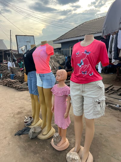 This image depicts an outdoor market scene in Oko Amakom, Nigeria. In the foreground, four mannequins are displayed, dressed in casual attire. The first mannequin from the left wears a black t-shirt and blue denim shorts. The second mannequin is dressed in a pink t-shirt and light blue denim shorts. The third mannequin, a child-sized mannequin, wears a red and white striped dress. The fourth mannequin, positioned on the right, is adorned with a bright pink t-shirt featuring a butterfly graphic and light beige cargo shorts. The mannequins are standing on circular bases, some of which are stabilized by rocks.
In the background, various market stalls are visible. To the left, stacks of what appear to be yams are piled high. A sign with the word "TIPHON" is mounted on a pole. Several people are present in the mid-ground, some standing and others seated. Clothing is displayed on racks in the background to the right. The ground is dusty and uneven, consistent with an outdoor market environment. The sky is overcast with scattered clouds.