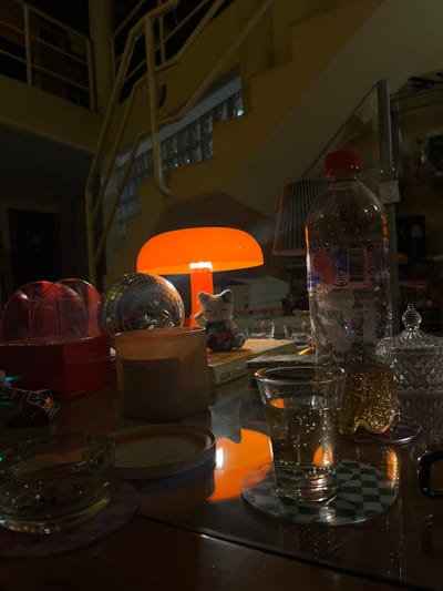 The image depicts a collection of objects on a reflective surface, possibly a glass table. These include a water bottle, glassware, a small lamp with an orange lampshade, a small figure, a disco ball, a candle, a jar, and a glass ashtray, against a blurred backdrop of what appears to be a multi-level interior with a staircase. The specific location within Santiago de Surco, Peru cannot be determined from the image.