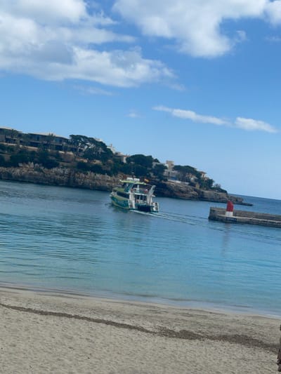 The image depicts a coastal scene within the bay of Porto Cristo, Spain, under a clear blue sky with scattered white clouds. In the foreground, a sandy beach occupies the lower third of the frame, showing light brown sand with a dark line of what appears to be seaweed or debris near the water's edge. The midground features calm, turquoise-blue water, disturbed by the wake of a green and yellow multi-deck passenger boat identified as "SEA PARADISE" visible on its hull. The boat is actively navigating towards the right side of the frame, moving away from the viewer, with a Spanish flag affixed to its stern. Indistinct human figures are visible on the boat's upper deck. To the right of the boat, a grey concrete pier extends into the water, culminating in a red and white striped lighthouse structure. Text "CLUB NAUTIC PORTO CRISTO" is legible on the side of this pier. In the background, a rocky cliff face rises from the water's edge, topped with green foliage and several multi-story buildings, likely residential or commercial structures. The horizon line of the open sea is visible beyond the pier on the right.
