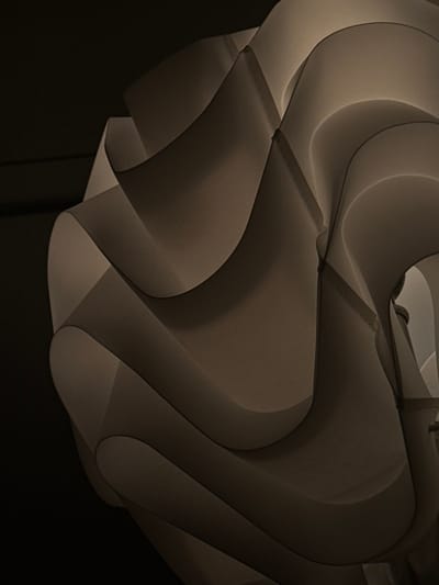 The image displays a close-up, abstract view of an illuminated lamp shade, likely situated indoors in the city of Hallandale Beach, United States. The lamp shade is constructed from multiple overlapping, curved, and undulating segments of a translucent, off-white or light beige material, possibly paper or plastic, creating a sculptural, organic form. An internal light source illuminates the material from within, causing brighter areas where the light is most direct and casting varied shades of brown and beige in the shadowed recesses and overlaps of the material, emphasizing its layered texture and three-dimensional shape. The background is uniformly dark, providing no additional environmental details, and there are no people, discernible objects other than the lamp, or actions depicted. The precise location within Hallandale Beach is not determinable from the visual content of the image.