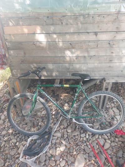 A green bicycle with a "STARLIGHT" logo is leaning against a wooden structure on a rocky ground. The bicycle has a black seat, handlebars, and tires. There are some discarded inner tubes on the ground near the bicycle. In the background, the wooden structure is weathered and appears to be part of an outdoor shelter or shed, covered with a translucent material at the top. The scene is illuminated by sunlight, casting shadows on the wooden structure. The location is identified as Korolevo, Ukraine.