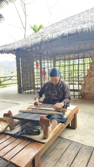An elderly woman, identified by traditional attire including a dark headscarf, dark long-sleeved shirt with light trim, and dark skirt, is depicted seated on a low wooden bench, engaged in weaving on a traditional loom. She wears two light-colored bangles on her left wrist and is barefoot, using her feet to brace part of the loom's tensioning mechanism. The loom features dark blue/black and white threads. A folded, multicolored woven fabric is placed on the bench between her feet.
The scene takes place under a shelter with a prominent thatched roof and rough wooden support poles. The background features a wall constructed from woven bamboo or thin wooden slats, with an exposed red lantern hanging within it. To the right, a section of an earthen or mud wall is visible. A concrete or paved floor is directly beneath the weaving area, transitioning to wooden planking in the immediate foreground. In the distant background, outside the structure, rolling hills or mountains with sparse vegetation are visible under an overcast sky. A person in a red garment is partially visible within the bamboo-walled section of the structure. The location is in Jiyang District, China.