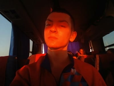 The image shows a young person with short, dark hair sitting on a bus or large vehicle. The subject is facing the camera with a serious expression. The interior of the bus has patterned seats, windows, and overhead storage. The lighting is warm and orange-toned, likely due to the time of day or artificial lighting within the vehicle. Another person can be seen in the background, seated behind the subject. The location of the photo is the inside of a bus in Ustinovo, Russia.