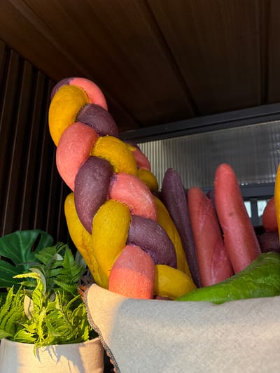 This is a close-up, eye-level, indoor shot of a display of vibrantly colored, braided bread loaves and some whole bread loaves in what appears to be a market stall or bakery in Phu Thuy, Vietnam. The bread is predominantly in shades of yellow, pink, and purple, with some darker red loaves also visible. A large, green, braided object, possibly a decorative element or another type of bread, is positioned in the foreground. Behind the bread, there are lush green artificial plants and a textured wooden background with vertical slats. The lighting suggests an artificial source, casting subtle shadows. There are no people visible in the frame.