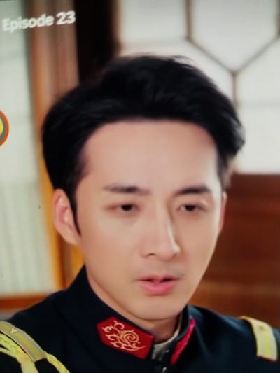 The image displays a close-up of a man's face and upper torso, with a text overlay indicating "Episode 23". The man has dark hair, fair skin, and is wearing a dark uniform with red and gold embellishments on the collar. The background is blurred, showing hints of wooden structures and possibly furniture, suggesting an indoor setting. There is no indication of the city of Palangka Raya, Indonesia, within the image itself.