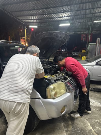 Two men are working on a car engine under the hood of the car. One man is wearing a red sweatshirt and black pants. He is bent over the engine, looking at it. The other man is wearing a white striped shirt and white pants. He is also bent over the engine, looking at it. The car is silver and black. The location is likely an auto repair shop, given the environment which appears to be indoors with industrial lighting. A sign for "KOSE" is visible in the background. The picture may have been taken in the city of Ula, Turkey, based on the user's request.