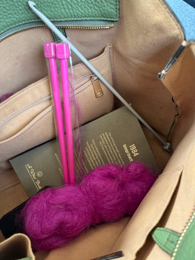 An overhead view inside an open bag in Elmbridge, United Kingdom, shows crafting supplies and a book. The bag's exterior is pale green and textured, with a visible zipper along a top edge. Its interior is lined with light tan material, including an internal zippered pocket featuring a gold-colored pull. Within the bag, two 8mm bright pink knitting needles are present in clear plastic packaging, positioned vertically in the center-left. A light grey crochet hook lies diagonally from the top-center-left to the bottom-right. A skein of fluffy magenta yarn is situated in the lower right. A dark-covered book, identified as "1984" by George Orwell, and bearing the imprint "A Wilco Book", is partially visible beneath the crafting tools. A small black object is partially visible beneath the yarn.