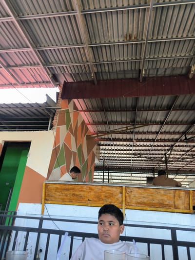 A medium, eye-level shot depicts a partially covered outdoor eating area in Matang, Indonesia. A young boy is seated at a table in the foreground, a white t-shirt visible. On the table are clear glasses with straws. In the background, two adults are visible behind a wooden barrier. One adult, wearing a white shirt, looks down, while the other, clad in brown, faces away. The eating area is roofed with corrugated metal sheets. The partially visible building structure features a geometric design in shades of brown, orange, and green, next to a green door.