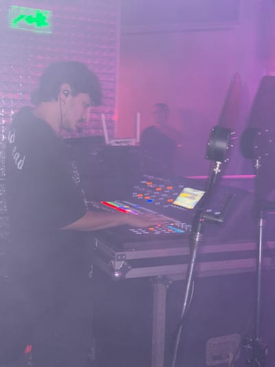 A person operates a complex electronic panel with multiple colored buttons and a small screen in a smoky, dimly lit setting. The person is wearing a dark shirt with white lettering, possibly "Dead." The scene includes stage lighting equipment and a metallic wall. An emergency exit sign is visible in the lower left corner.