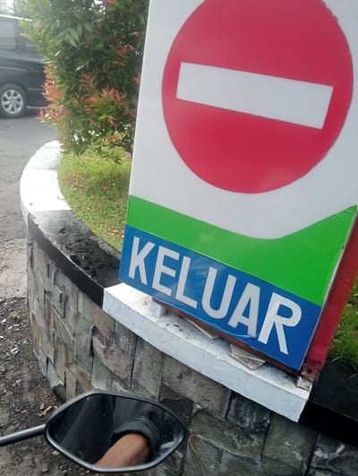 In Surabaya, East Java, an image captures a close-up of a red and white "no entry" sign and a blue sign with white lettering that reads "KELUAR," which translates to "EXIT" in Indonesian. The signs are mounted on a white base, which rests on a stone wall. To the left of the signs, a black vehicle is partially visible parked on a street, and lush green grass and foliage surround the area. The reflection in a motorcycle's rearview mirror shows a person's arm wearing a black shirt with rolled-up sleeves. The overall scene depicts an outdoor setting, likely in a public area or establishment, with clear signage indicating directions.