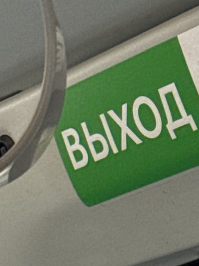 This is a close-up, angled shot of a green sign with white text that reads "ВЫХОД" in Cyrillic, meaning "EXIT". The sign is positioned diagonally and appears to be on a metallic surface, possibly a door or a piece of machinery, given the curved metallic object in the foreground which could be a handle or part of a railing. The background is a textured, light gray surface. The lighting is even, suggesting an indoor setting or an overcast day. The image focuses tightly on the text and the surrounding material, giving no broader context of the scene or location beyond what is implied by the text itself.