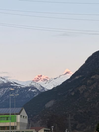 This image depicts a mountainous landscape in Lalden, Switzerland, during either sunrise or sunset. The sky transitions from light blue at the top to a pale pink-orange closer to the horizon. Four thin, dark horizontal power lines traverse the upper sky, with a faint, elongated cloud situated below the lowest line.

In the mid-ground, two prominent snow-capped mountain peaks are visible, their summits intensely illuminated with a pink-orange glow from the low sun. Surrounding these peaks are other snow-covered mountains, appearing in muted light or shadow. A large, dark, steep, tree-covered mountain slope occupies the right side of the frame, extending from the mid-section downwards, mostly appearing in shadow. Another dark, tree-covered slope is partially visible behind the building in the foreground on the left.

In the left foreground, a portion of a multi-story building is visible. Its light-colored roof is equipped with dark solar panels. The top visible floor has a white facade with three small rectangular windows. Below this, a section of the building features a vibrant green facade with multiple horizontal windowpanes. An external staircase or similar structure is visible adjacent to the green section. Two dark, double-headed streetlights are positioned in front of this building. In the lower right, a dark, industrial-style pylon or power pole is partially visible, accompanied by several smaller, single-headed streetlights along the bottom edge. No individuals or animals are present in the scene.