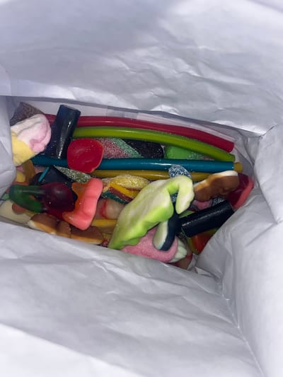 An overhead view inside a white paper bag reveals a jumble of colorful, gummy candies. The candies are a mix of shapes and sizes, including long sticks of red, green, and blue, as well as smaller, more irregularly shaped pieces. Some of the candies are coated in sugar. The bag is filled to the brim with the confectionery. The image does not depict any location in Stary Chultem, Russia, nor does it include any people or actions.