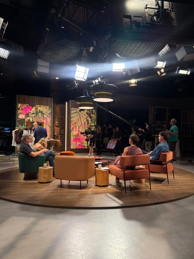 The image depicts a television studio located in Sant Joan Despí, Spain. In the foreground, four individuals are seated on a circular wooden platform. Two individuals, a man and a woman, are seated in chairs facing each other and the camera. The man is wearing a dark shirt and glasses, and the woman is wearing a patterned top and jeans. Another man, wearing a blue denim jacket, is seated to the right of the woman, facing her. To the left of the man in the blue jacket, a woman in a maroon top is seated, facing the man in the blue jacket. The set features large, vibrant floral backdrop panels and studio lighting equipment suspended from the ceiling. Several crew members are visible in the background, operating cameras and other production equipment, indicating an active filming or broadcast.