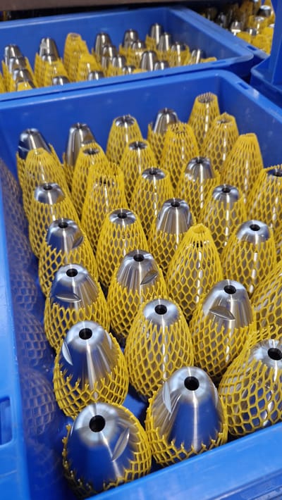 The image shows a close-up, eye-level view of multiple metal objects, likely machine parts, organized within blue plastic containers. These objects are conical in shape, with a hollow opening at the top. Each metal part is encased in a yellow, netting-like protective material. The objects are arranged in multiple rows and appear to be in a state of storage or transport. The background suggests an industrial setting, possibly a factory or warehouse, with more containers of similar items visible. The lighting is bright and even, highlighting the metallic sheen of the parts and the texture of the yellow netting. The specific location of Ichenhausen, Germany, is not visually discernible from the image itself, which focuses solely on the product and its packaging.