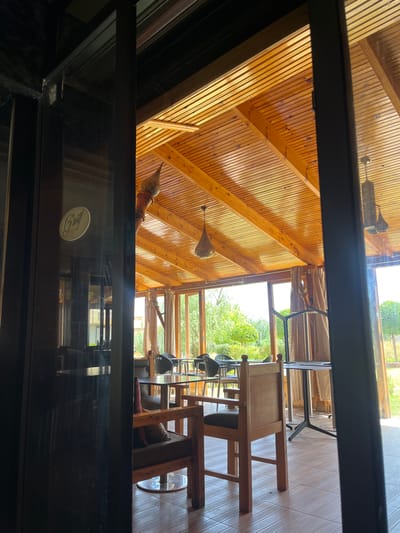 The image shows the interior of a restaurant with a wooden ceiling and large windows looking out onto a garden. Several tables and chairs are visible, as well as decorative hanging lamps. The overall atmosphere appears to be relaxed and inviting. A logo with the text "Gsouif" is visible on the glass door in the foreground. The scene is situated in El Hajeb, Morocco.