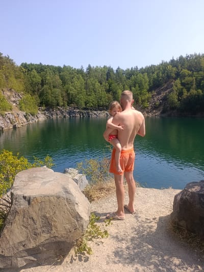 A father stands on a rocky shore, holding his young daughter. He is wearing orange swim trunks, and his daughter is in a red and white patterned swimsuit. The lake before them is a deep emerald green, reflecting the lush green trees that line the opposite shore. Rocky cliffs rise behind the trees, suggesting a quarry or a natural rock formation. The sun is high in the sky, casting sharp shadows and indicating it is midday. The weather appears to be warm and clear, perfect for a swim. The scene evokes a sense of peace and family enjoyment in a natural setting. There is no visible text in the image.