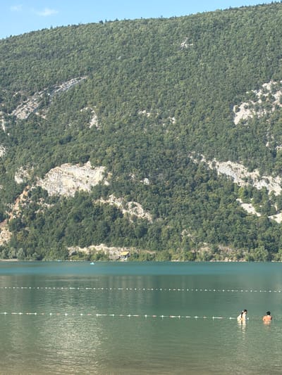 The image depicts a scenic view of the Lac d'Aiguebelette in Novalaise, France. A steep, tree-covered mountainside dominates the background, with a prominent rock face exposed partway up. In the foreground, a serene, green lake is visible, marked by two parallel lines of buoys. Two individuals are in the water, with one person partially submerged and facing towards the viewer, while another person stands further back, their back to the camera. A small building with a yellow roof is situated on the far bank of the lake, nestled amongst trees. The sky is a clear, pale blue with a few wispy clouds.