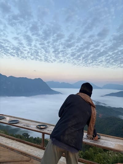 A person stands on a wooden structure, gazing at a mountain range veiled in mist and clouds. The sky is filled with puffy, textured clouds. The individual wears a dark jacket, a scarf, a hat, and light-colored pants. A wooden table with objects on it is visible on the structure. The overall scene is serene and scenic.