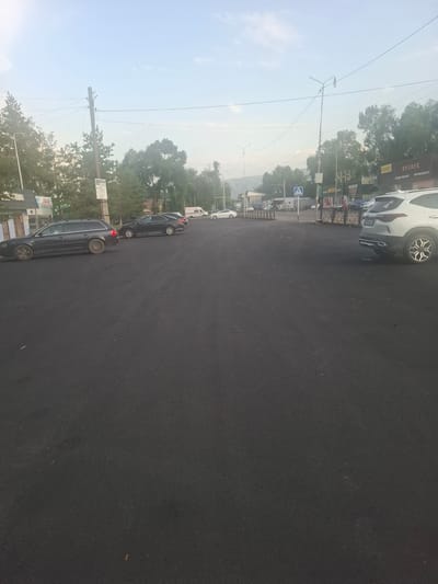 In Uzynagash, Kazakhstan, an outdoor scene captured from a slightly elevated perspective reveals a newly paved asphalt parking lot. The lot is populated by several parked vehicles, including a dark-colored station wagon on the left, a black sedan in the middle, and a white SUV on the right. In the background, a low-rise commercial building with several storefronts is visible, featuring signs such as "RYSBEK," "shagymmarket," and "МИНИМАРКЕТ." Adjacent to the building are trees and utility poles, with power lines crisscrossing the sky. The sky itself is a pale blue with some haze, suggesting it might be early morning or late afternoon. The overall impression is that of a functional commercial area in a semi-urban or suburban setting.
