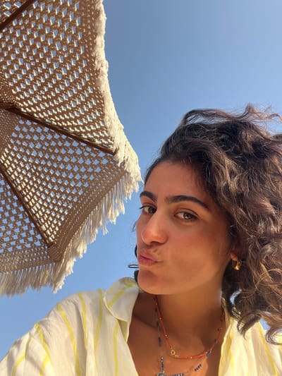A low-angle, close-up photograph depicts a female subject in the city of Aljezur, Portugal, positioned against a clear blue sky. The subject, occupying the right half and lower portion of the frame, has dark, curly hair and dark eyes. She is looking directly at the viewer with her lips puckered. She wears a light-colored, collared shirt with thin yellow vertical stripes, a small gold hoop earring on her visible right ear, and multiple necklaces including a red beaded strand and a longer dark blue beaded chain. In the upper left quadrant, a large, light-beige, open-weave parasol with a distinct fringed border is partially visible, providing shade.