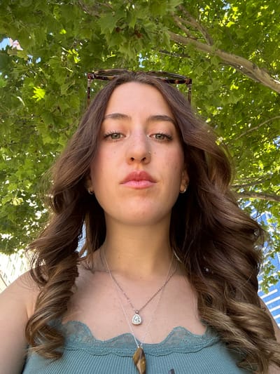 A young woman with long, wavy brown hair poses outside. She wears a blue-green top and two delicate necklaces, one with a heart-shaped pendant and the other with a larger, brown stone. Sunglasses perch atop her head. Green trees fill the background, offering dappled sunlight. Her fair skin is accented with freckles. Her eyes are green, and she wears subtle makeup.