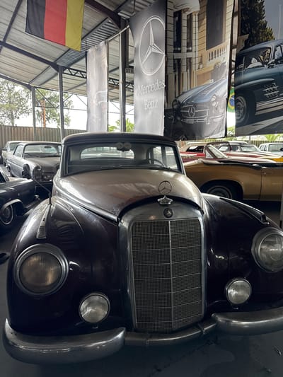 The image depicts a collection of classic Mercedes-Benz cars inside a large building, likely a garage or showroom. The primary subject is a dark-colored vintage Mercedes, with a prominent grille and rounded headlights. Behind it, other classic cars are visible, some of which are tan or beige. There are also hanging banners featuring Mercedes-Benz logos and images of classic car models. A German flag hangs from the building's ceiling. The location appears to be a covered structure with corrugated metal walls and a steel framework. While the specific location in Kraaifontein, South Africa, is not visually identifiable, the image suggests an automotive museum or a specialized vintage car collection facility. There are no people visible in the shot.