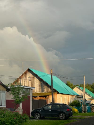 The image captures a rural scene in Yakshur-Bod'ya, Russia, under a cloudy sky with a faint rainbow. A black SUV is parked on a gravel area in front of several wooden houses. One house, in the foreground, has a light-colored wooden facade and a vibrant teal roof. Another house to the right also has a teal roof, and a third, partially visible house on the far left, has a beige facade with a dark red lower section. Power lines crisscross the sky above the houses, supported by wooden poles. A tree with green foliage stands between two of the houses. The ground is covered with grass, some flowering bushes, and gravel. The lighting suggests it might be late afternoon or early evening, with sunlight illuminating the wooden buildings from the side.