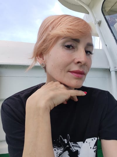 A woman with peach-colored hair, wearing a black t-shirt and heart-shaped earrings, rests her chin on her hand. She has on bright red nail polish. The background appears to be the exterior of a boat or ferry, with white railings and a cloudy sky visible. The image was taken outdoors.