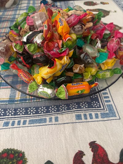 A close-up, overhead shot shows a clear glass bowl overflowing with a variety of individually wrapped candies. The candies are in brightly colored wrappers, predominantly red, orange, yellow, green, and pink, with some featuring gold or silver foil accents. Many of the wrappers have text and images indicating flavors such as "Toffee," "Dolci Note," "Menta Everest," and "Caffè Latte." The bowl sits on a tablecloth with a blue and white checkered pattern and a floral border. In the background, a portion of a dark surface is visible, and further out, a glimpse of what might be a table or other furniture. The scene is indoors, likely in a domestic setting.