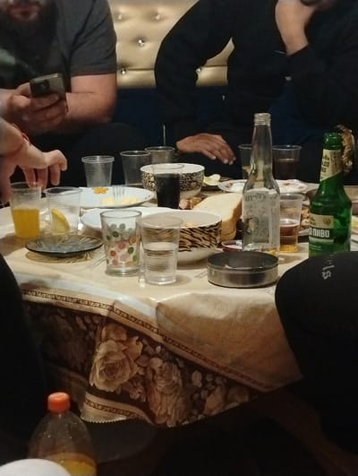 The image depicts an indoor social gathering around a table in the city of Byal Izvor, Bulgaria. Four individuals are partially visible. On the left, a person wearing a dark grey t-shirt and beard holds a smartphone. To their right, another person in a black long-sleeved shirt sits with their left hand resting. A third person's hand reaches into the frame from the far left, and a fourth person's knee is visible in the bottom right.

The table is covered with a light-colored tablecloth adorned with a brown and gold floral pattern featuring roses. Numerous items are arranged on the table: a glass of orange juice with a lemon slice, multiple clear plastic cups (some empty, some containing dark or light brown liquids), two clear glasses of water (one with polka dots), slices of white bread, several white ceramic plates (some with food remnants), two black and white patterned ceramic bowls containing unidentifiable food, an empty clear glass bottle, a green glass beer bottle labeled "ПИВО" in Cyrillic, and a dark metal ashtray. A plastic bottle with an orange cap and orange liquid is in the lower left foreground. The background features dark blue upholstered seating with metallic studs, set against a light-colored, possibly padded or upholstered wall section also adorned with metallic studs.