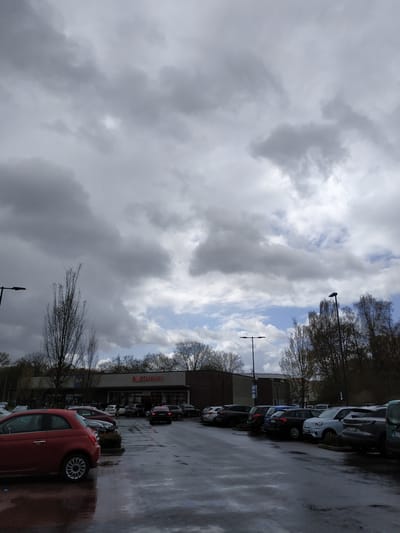 The image shows a parking lot outside a Rossmann store in Lünen, Germany on a cloudy day. Parked cars are visible, including a red Fiat 500 in the foreground. The parking lot surface is wet. Bare trees are present between the parking lot and the building. The Rossmann store has signage visible. A street lamp is also visible.