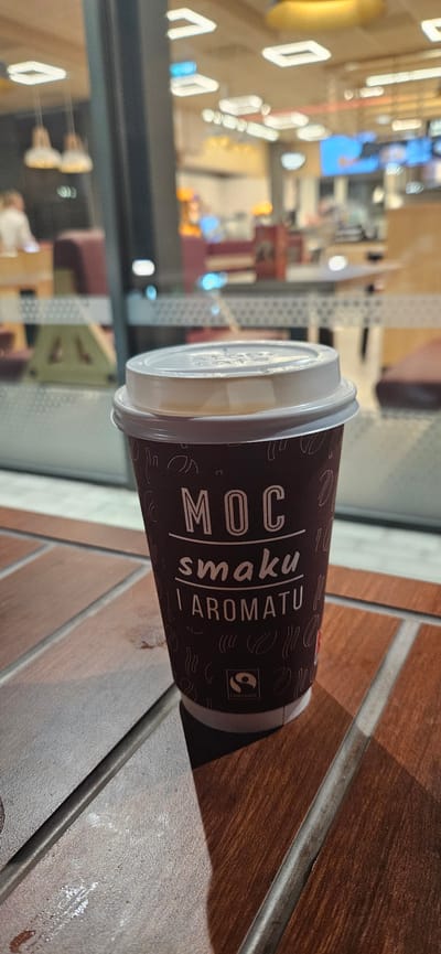 A close-up, slightly angled shot shows a disposable coffee cup with a white lid, placed on a wooden table. The cup is dark brown and has white text that reads "MOC smaku I aromatu". Below this text, there is a small Fairtrade logo. The background is blurred and appears to be an indoor setting, possibly a cafe or restaurant, with reflections of lights and architectural elements on a glass window. The overall lighting suggests it might be evening or a dimly lit interior.