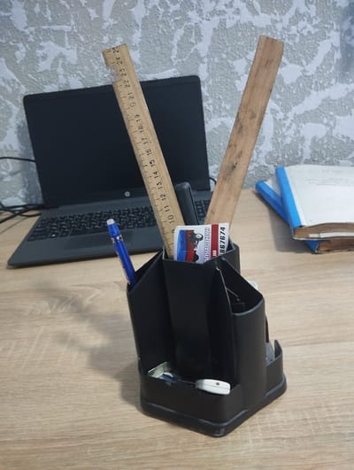 The image shows a desk with a laptop, a pen holder, rulers, a business card, and books. The desk is made of wood and has a light wood grain pattern. The laptop is black and is open, with the screen facing forward. The pen holder is black and is filled with various writing instruments and a business card. One ruler is made of wood and is placed diagonally in the pen holder. Another ruler is made of wood and is also in the pen holder. The business card has an image of a vehicle on it and some text. Two books are stacked on the right side of the desk, with blue covers. The background is a textured wallpaper. The scene is indoors, likely an office or study. No specific location in Vinkivtsi, Ukraine, can be determined from the image.