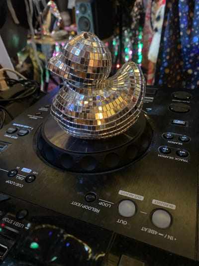 The image shows a DJ controller with a heart-shaped disco ball resting on the turntable. The controller is black and has various buttons and knobs. Text visible on the controller includes "TRACK SEARCH", "LOOP", "USB1 STOP", "CUE/LOOP", "CALL", "BEAT SYNC", and "TEMPO". The disco ball is made of small mirrored squares, reflecting light. The background is cluttered with various objects including speaker, LED lights, cloth and cables. The image does not contain information to verify it was taken in Baan Nai Suan, Thailand.
