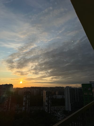 The image shows a sunset over a densely populated urban area, likely a residential district. The sun, a bright orange disc, is low on the horizon, partially obscured by clouds. The sky is a gradient of colors, from pale blue at the top to warm oranges and yellows near the horizon, with dramatic, textured clouds in shades of gray and white.
Below the sky is a panorama of buildings, predominantly high-rise apartment blocks. Some are sleek and modern with dark facades and numerous windows, while others appear to be older or of a different architectural style. The buildings vary in height and density, forming a cityscape that stretches across the frame. In the foreground, parts of a balcony railing are visible, suggesting the photo was taken from a high vantage point, such as an apartment window or a balcony. Trees and darker, less defined structures are visible between and around the buildings, indicating the presence of parks or lower-rise areas. A parking lot with numerous cars is visible between some of the buildings. The overall impression is one of an urban landscape at dusk, with the natural beauty of the sunset contrasting with the man-made environment.
The location specified, Feklistovo, Russia, is a village in the Vologda Oblast, which is a rural area. The scene depicted in the image is clearly an urban environment with high-rise buildings, which is not consistent with the known characteristics of Feklistovo. Therefore, the provided location information is likely inaccurate for this image.