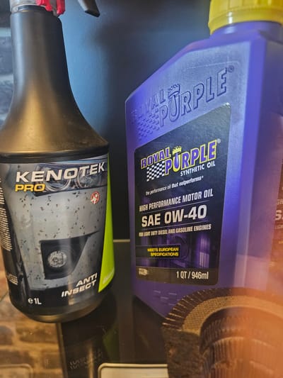 The image displays two bottles of automotive fluids placed on a surface, possibly a shelf or table. On the left is a black spray bottle with a lime green label featuring the brand "KENOTEK PRO" and text "ANTI INSECT" along with "e1L". The bottle has some smudges and marks on its surface. To the right is a purple bottle with a yellow cap, labeled "ROYAL PURPLE SYNTHETIC OIL". This bottle contains "HIGH PERFORMANCE MOTOR OIL SAE 0W-40 FOR LIGHT DUTY DIESEL AND GASOLINE ENGINES" and also states "MEETS EUROPEAN SPECIFICATIONS" and "1 QT / 946ml". A small portion of a cog or gear-like object is visible in the foreground, out of focus. The background consists of a brick-patterned wall and a darker, possibly blue, wall section. The scene appears to be an indoor setting. There are no people or animals visible in the image. No specific location in Bemmel, Netherlands can be identified from the image.