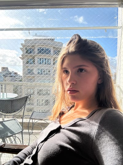The image depicts a young female, illuminated by natural sunlight, seated on a high-rise balcony or terrace in the Mont'Serrat neighborhood of Porto Alegre, Brazil. She is oriented towards the right side of the frame, with her gaze directed upwards and to the right, beyond the visible area. Her long, light brown hair is distinctly highlighted by the sun, which casts shadows across her face. She wears a dark grey, ribbed, collared top with buttons and a thin gold necklace. A small stud earring is visible in her right ear.

The balcony features outdoor furniture; specifically, a dark, possibly metallic, table and two chairs are visible to the left, partially obscured. The entire outdoor perimeter is enclosed by a white, grid-patterned safety net. In the background, beyond the net, a distinctive multi-story building identified as Edifício San Remo (Rua Quintino Bocaiúva, 1546) is prominently visible, characterized by its light-colored facade and tiered architectural design. Other urban structures are present in the distant background. The sky is bright blue with scattered white clouds, indicating clear weather.