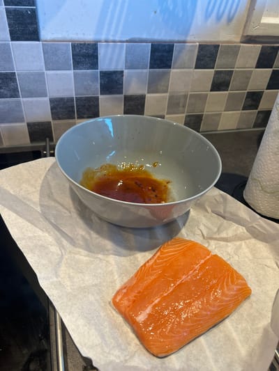 A raw salmon fillet lies on white parchment paper on a dark countertop. Next to it, a light gray bowl contains an orange sauce with red pepper flakes. In the background, a black and white tiled backsplash is visible, along with a roll of paper towels. The scene appears to be in a kitchen setting.