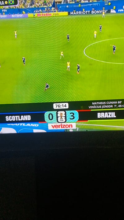 A soccer match is being broadcast on a screen. The score is Scotland 0, Brazil 3. The time is 76:14. The game appears to be taking place in a stadium with a bright green field and yellow seating in the background. There are several players on the field, wearing dark blue and yellow uniforms. A Verizon logo is visible at the bottom of the screen. The broadcast also shows a ticker with player names and times, indicating that Matheus Cunha scored at 60' and Vinícius Júnior scored at 7' and 45+3'. The image is taken from a slightly angled perspective, with the bottom portion of the screen showing a dark, textured surface, possibly a television stand or furniture. The location is identified as Pistanmazor, Tajikistan, however, this is not verifiable from the image content itself.