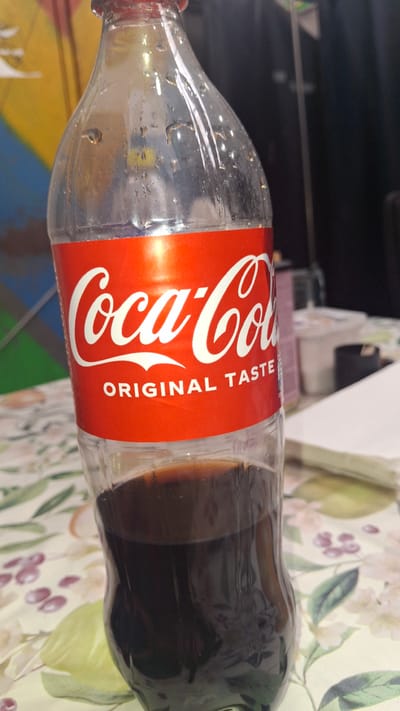 A clear plastic bottle of Coca-Cola, with condensation on the outside, is shown at eye-level. The label, red with white lettering, prominently displays "Coca-Cola ORIGINAL TASTE." The bottle is partially filled with dark brown liquid, and it rests on a table with a floral-patterned tablecloth. In the background, out of focus, are some containers and a dark curtain. The image does not provide any information that would allow for the determination of its location in the city of Račice, Czechia.