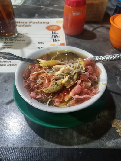 This close-up image captures a meal served on a dark, textured table, likely at a casual eatery during the evening or night, indicated by the dim artificial lighting. The central focus is a white bowl containing a soup-based dish, identified by visible menu text as "Miso Padang."
The dish features thin white noodles, shredded white meat (likely chicken), bean sprouts, and distinctive bright pink crackers (kerupuk) as a topping. Two decorative metal spoons are placed in the bowl. The bowl rests on a green saucer.
To the left of the bowl, a tall glass of iced tea with a straw is partially visible. Behind it, a menu lists food items and prices, with "Miso Padang" clearly legible. Other visible items include "ayam" (chicken), "sapi" (beef), and "ceker" (chicken feet) priced at "16K," "18K," and "16K" respectively, with "K" likely denoting thousands of Indonesian Rupiah. Additional partial text includes "Opak," "Telor" (egg), "kowut," and "apa utuh."
On the right side of the frame, an orange squeeze bottle, possibly containing chili sauce or sambal, a small woven basket, and another orange lidded container are visible. No people are present in the image. The scene depicts a typical local dining experience.