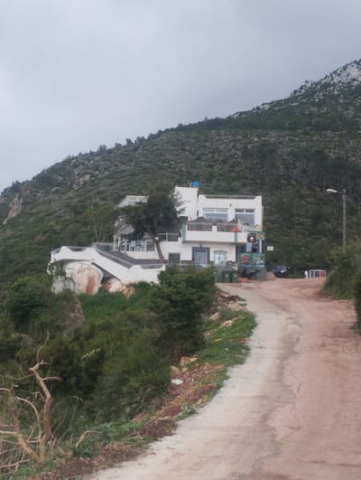 The image depicts a building situated on a rocky outcropping along a dirt road in Tétouan, Morocco. The building, predominantly white with multiple levels and balconies, seems to be a café or restaurant given the signage visible on its facade. It features a staircase leading down from the top level. The building's foundation is partially supported by a large rock formation. Lush green vegetation covers the slope leading up to the building and spills onto the roadside. A mountain rises in the background, covered with trees and shrubs. The sky is overcast, providing diffused lighting. A street lamp is visible on the right side of the road. Some vehicles are parked near the building, suggesting customer traffic. A few trash bins stand near the building's entrance.