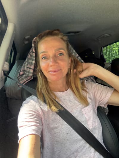 A woman with blonde hair, wearing a light pink t-shirt and a seatbelt, is in the driver's seat of a car. She is smiling and touching her hair. The car is packed with luggage and blankets, suggesting a road trip or moving. Outside the car window, trees are visible, indicating a rural or forested area. The image was taken in Portovoe, Russia.