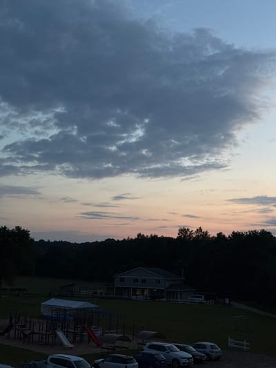 The image depicts an outdoor scene at dusk. The sky is a gradient of blues and pinks, with dark clouds scattered across the upper portion and lighter clouds transitioning to the horizon. A line of dark trees forms the backdrop, with a hill rising behind them.

In the middle ground, a two-story building with multiple windows is visible. A white van is parked in front of it. A children's playground with slides and swings is in the foreground, partially covered by a blue and white tent. Several cars are parked in a gravel lot in the lower foreground.

The scene appears to be set in a rural or suburban area. The location in Ratnieki, Latvia is not evident from the visual information in the image. There are no people visible in the image, nor any interactions occurring. The overall atmosphere is calm and serene, characteristic of twilight.